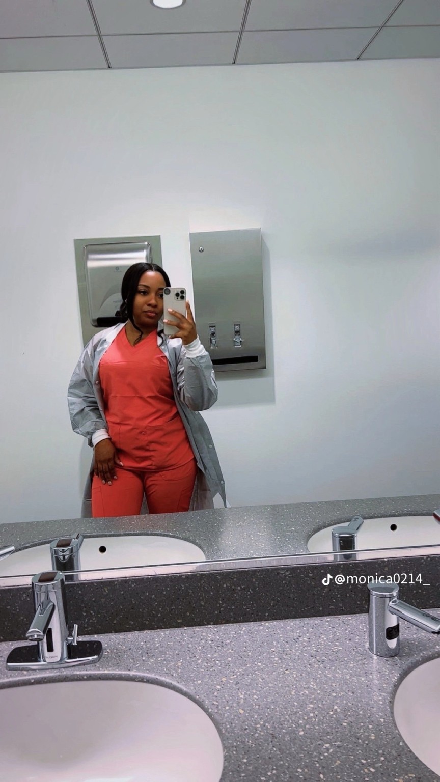 Monica Ross taking a mirror selfie in coral scrubs and a light grey jacket